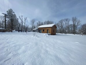 Una cabana de fusta amb un teulat cobert de neu, envoltada per un paisatge nevós i arbres sense fulles.
