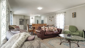 A living room with a brown leather sofa, a green armchair, and a glass coffee table. There is a dining area with wicker furniture and a kitchen with a refrigerator.