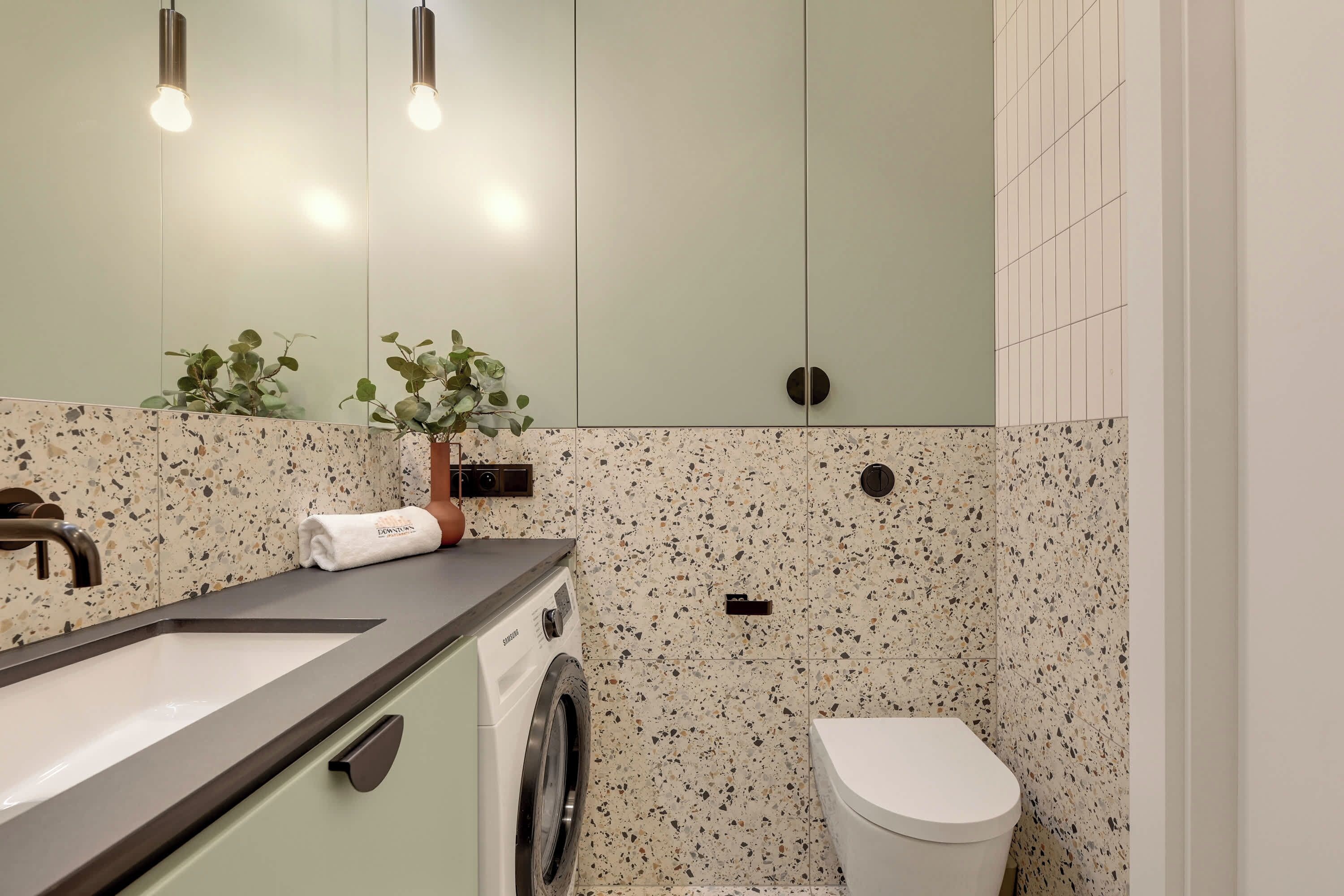 A bathroom with a sink, mirror, towel, washing machine, toilet, and a plant.