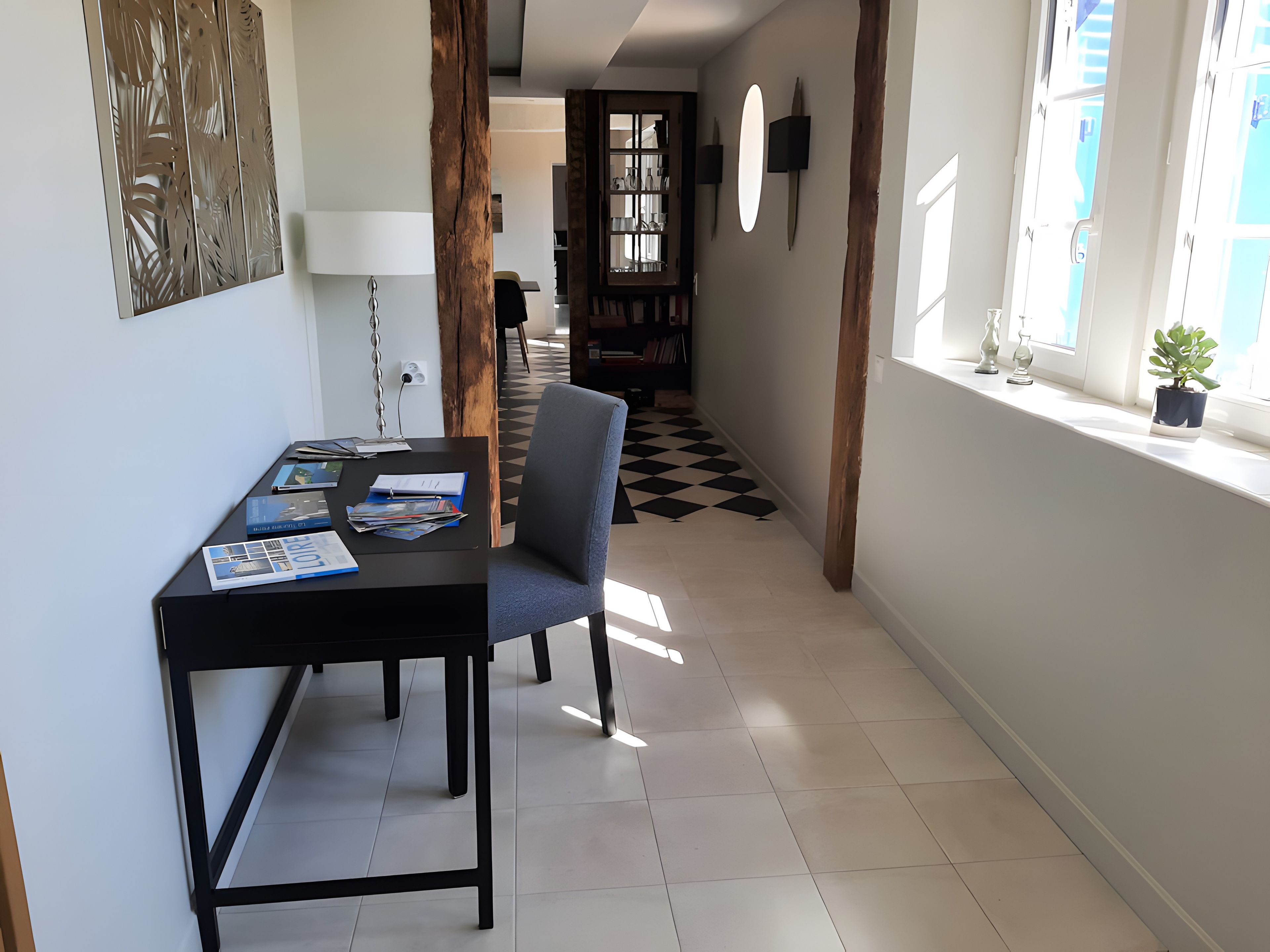 Un coin repas avec une table noire, une chaise bleue et une chaise noire. Le sol est carrelé en noir et blanc, il y a une porte en bois et une fenêtre avec une plante.