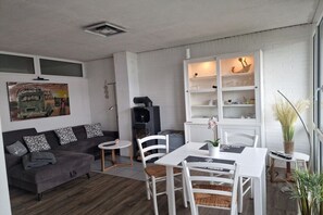 A compact living space with a sofa, dining table, chairs, a small kitchen area, and a bookshelf. - Holiday home with 2 bedrooms (Butjadingen)