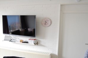 A flat-screen TV mounted on a white brick wall, a clock, and a radiator below. - Holiday home with 2 bedrooms (Butjadingen)