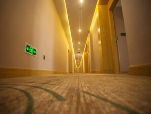 A hotel corridor with a carpeted floor, a green emergency exit sign, and a wooden door on the right.