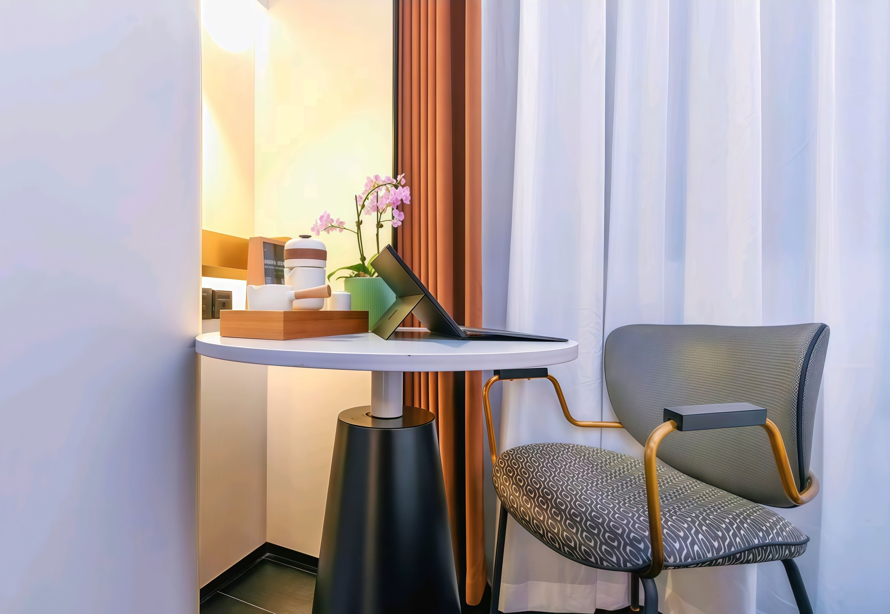 A hotel room with a small round table, a chair, a potted orchid, and a wooden side table with a laptop and a box.