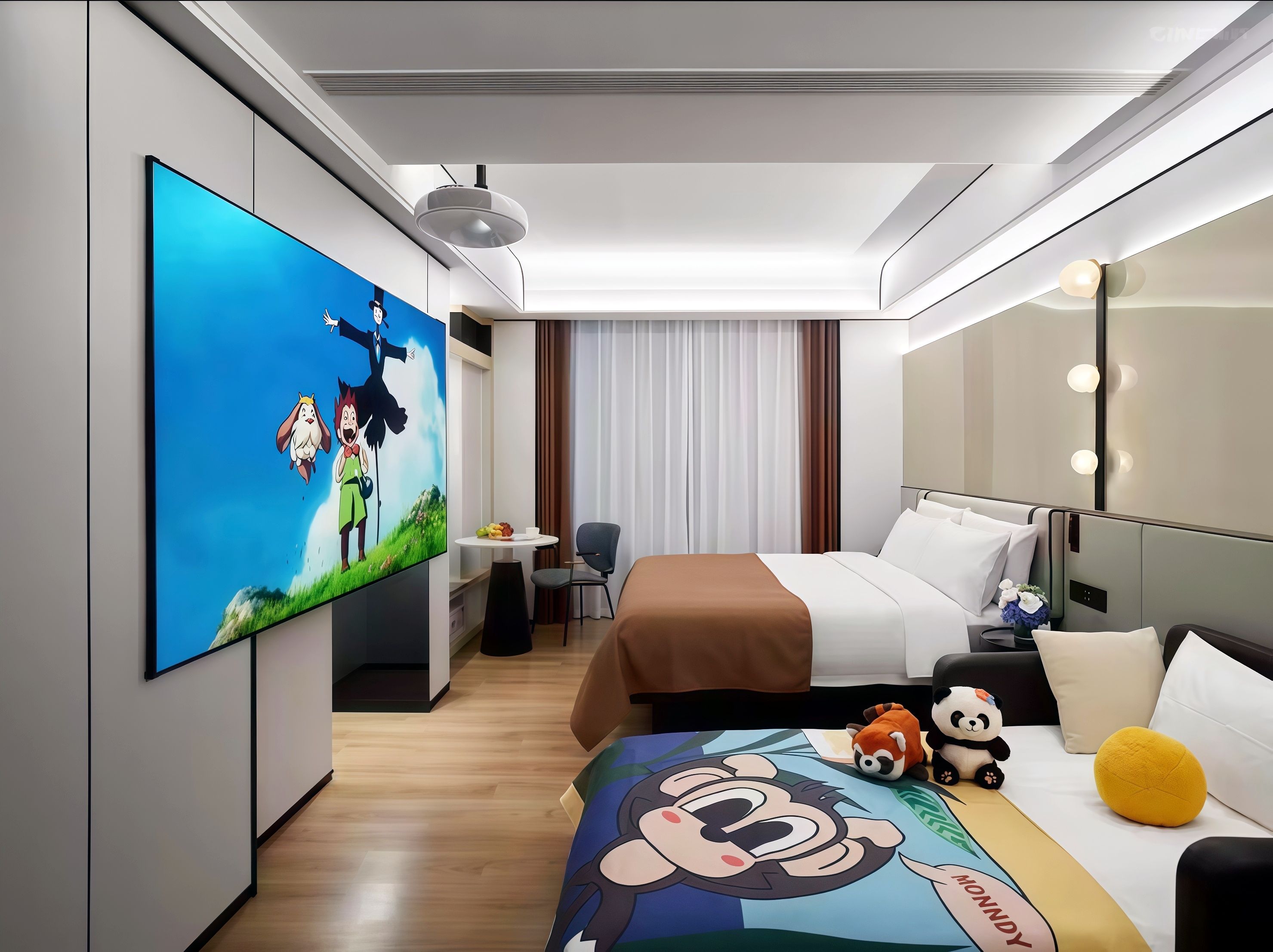 A hotel room with a large flat-screen TV, a bed with white and brown bedding, a small table with fruit, a chair, and plush toys on the bed.