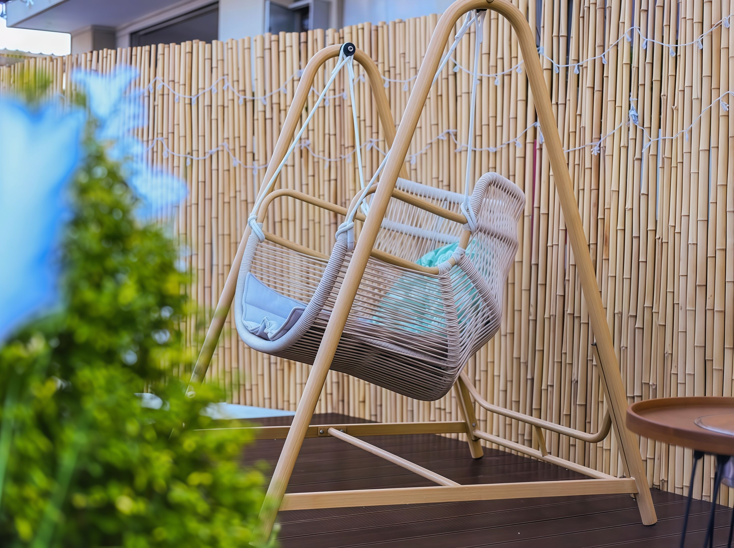 A wicker swing chair with a cushion, set against a bamboo fence backdrop.
