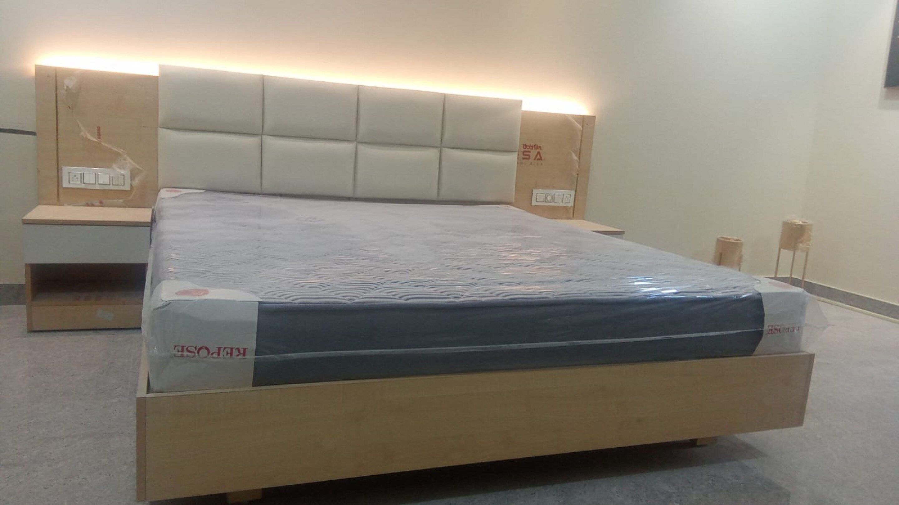 Un letto con testiera imbottita, un comodino in legno e un materasso in una stanza dalle pareti spoglie.