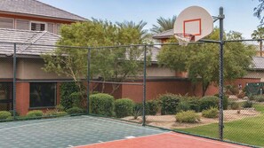 Ein Basketballplatz im Freien mit einem Maschendrahtzaun, umgeben von Grünflächen und einem Gebäude mit rotem Dach.