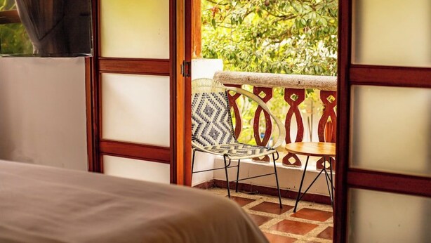A room with a bed, a small table, and a chair with a patterned cushion. A balcony with a wooden railing and a view of greenery is visible. - Hotel Portales del Tayrona by DOT Tradition (Santa Marta)