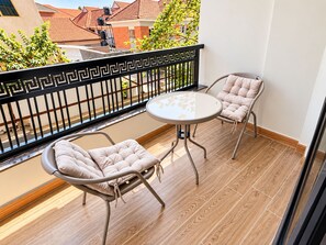 A balcony with a round table and two cushioned chairs, a decorative black railing, and a view of buildings and trees. - Advanced Homes (Kampala)