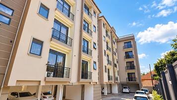 A multi-story residential building with balconies, a paved driveway, and parked cars.