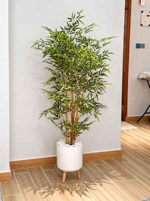 Un elegante pedestal cilíndrico blanco sostiene una planta de bambú, ubicado frente a una pared lisa con piso de madera.