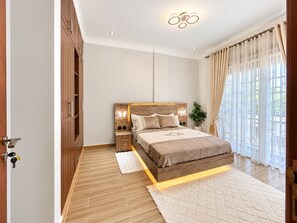 A bedroom with a wooden headboard, a bed with a quilted cover, a nightstand, a wall-mounted light, and a window with curtains. - Advanced Homes (Kampala)