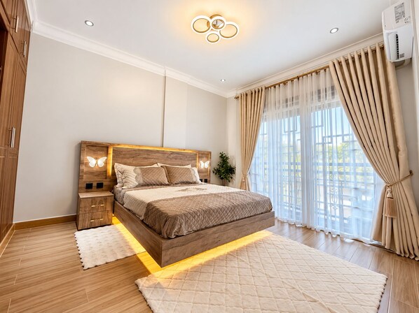 Un dormitorio moderno con una cama de madera, mesitas de noche, un amplio ventanal con cortinas y una lámpara en el techo.