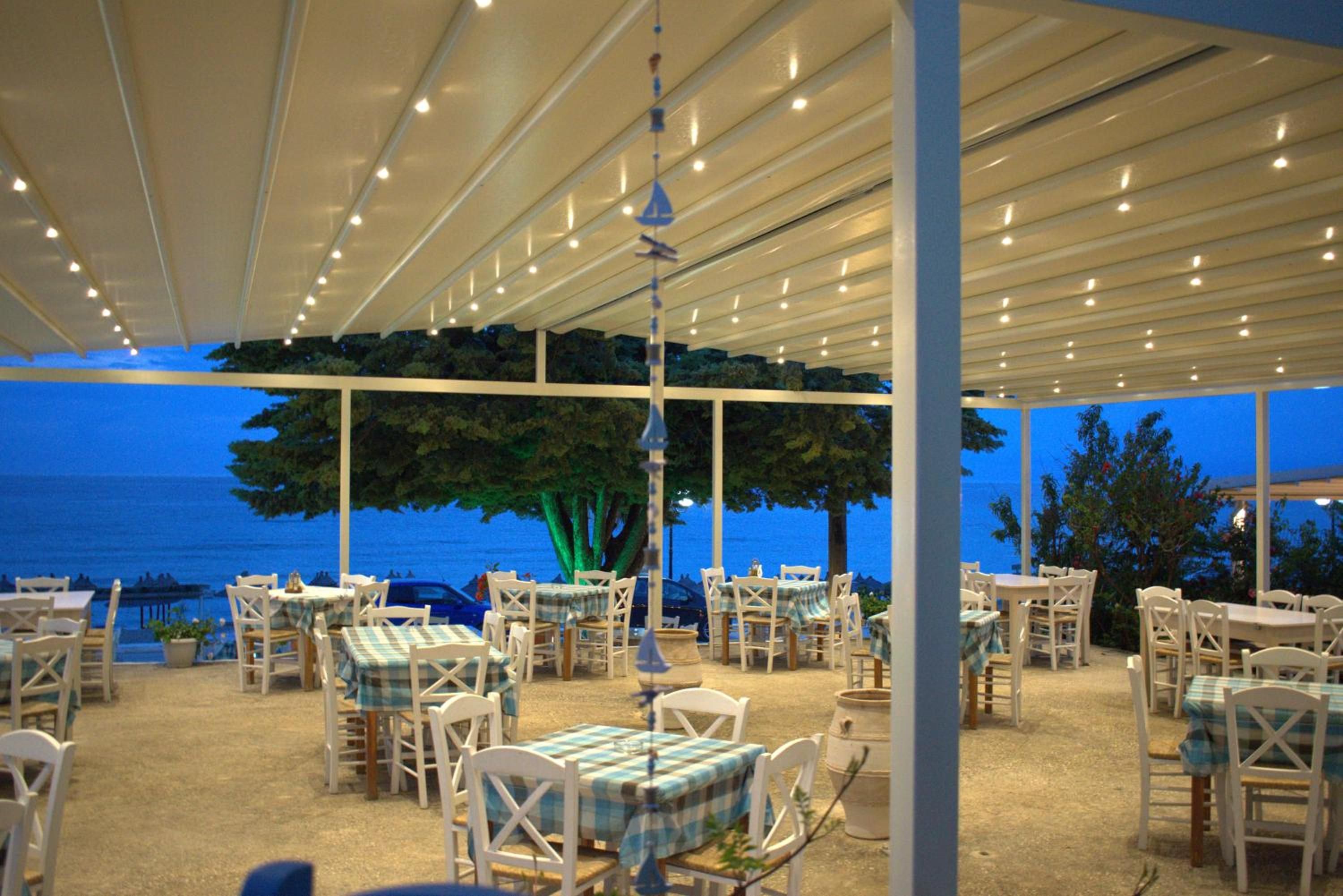 Ein Essbereich im Freien mit Tischen und Stühlen, einer Pergola darüber und Blick aufs Meer.