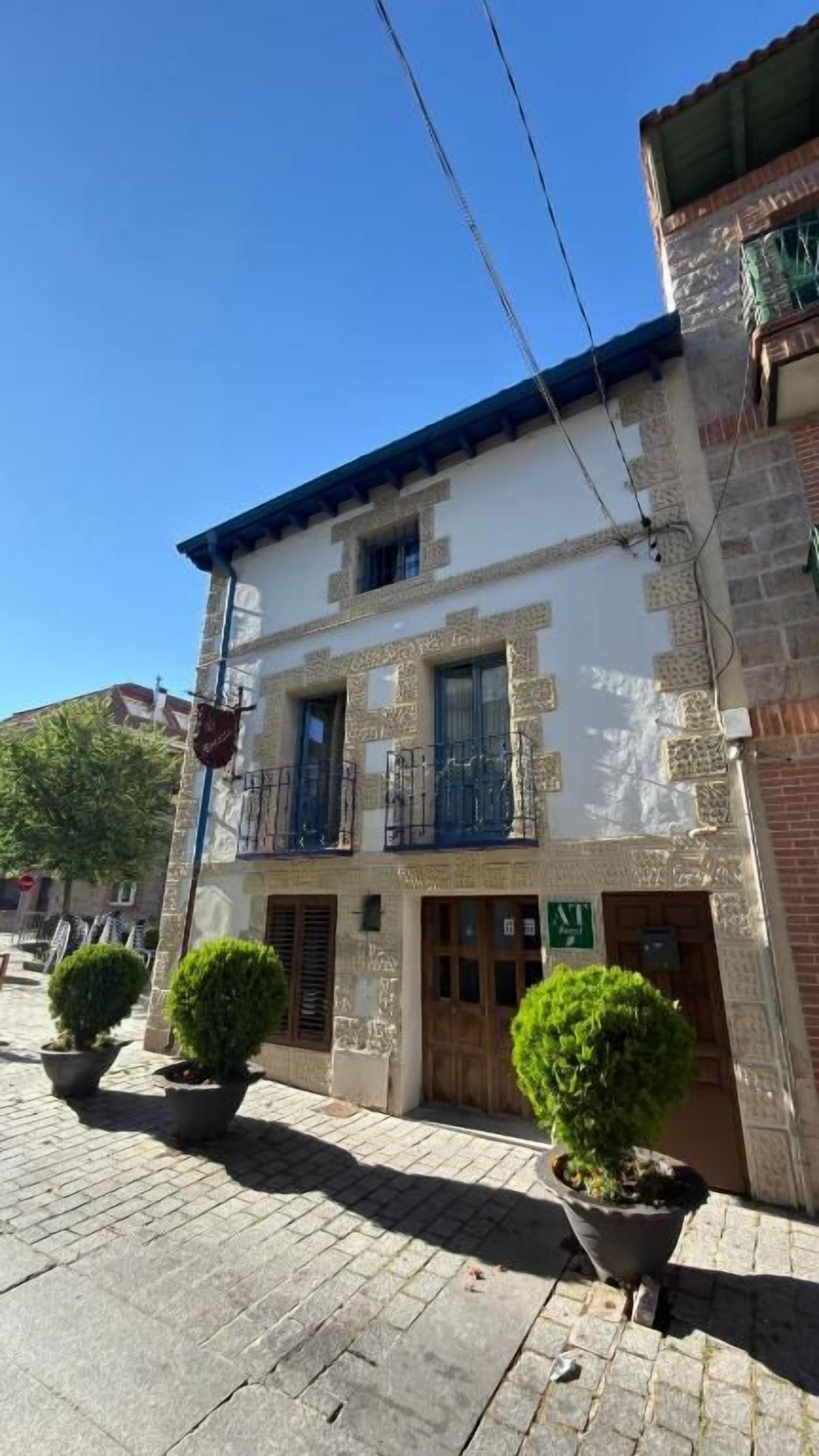 Un edificio de dos pisos con balcón, plantas en macetas y una puerta con un letrero.