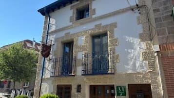 Un edificio de dos pisos con balcón, plantas en macetas y una puerta con un letrero.