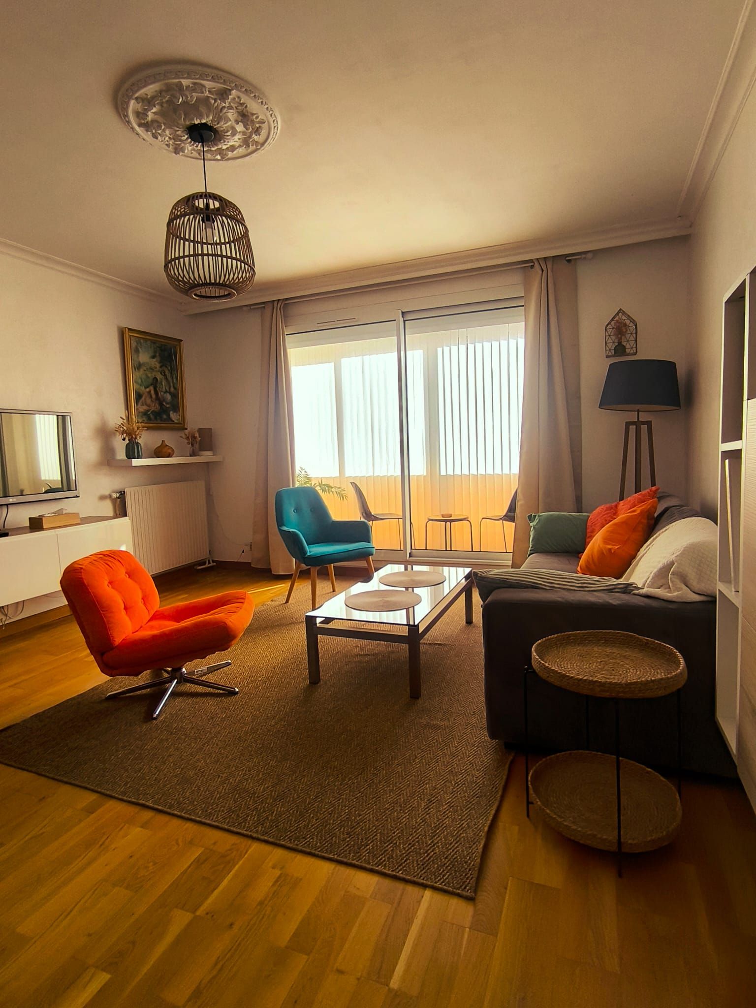 Een woonkamer met een bank, fauteuils, een salontafel, een televisie en een groot raam met gordijnen.