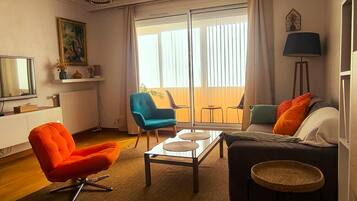 Een woonkamer met een bank, fauteuils, een salontafel, een televisie en een groot raam met gordijnen.