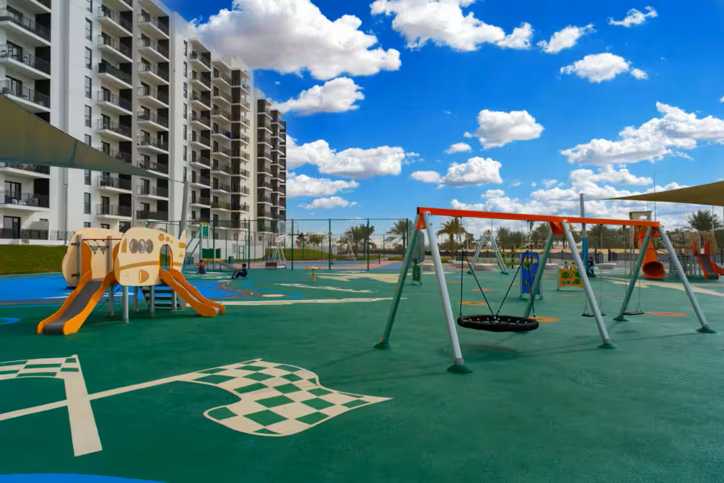 Ein Spielplatz mit Schaukeln und Rutschen, umgeben von einer grünen Wiese und einem mehrstöckigen Gebäude.