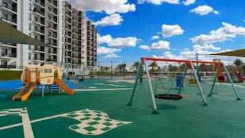 Ein Spielplatz mit Schaukeln und Rutschen, umgeben von einer grünen Wiese und einem mehrstöckigen Gebäude.