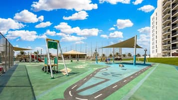 Un parque infantil con suelo verde, una estructura de juegos amarilla y un edificio grande al fondo.