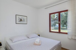 Un lit simple avec une couvre-lit et des oreillers à motifs floraux, un tableau encadré au mur et une fenêtre donnant sur des arbres et de l’herbe.
