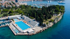 Vue aérienne d’une zone côtière avec une grande piscine, un port de plaisance et un paysage urbain en arrière-plan.