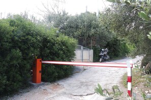 Une entrée fermée par une barrière rouge et blanche, entourée d’une végétation luxuriante et un sentier menant à un portail.