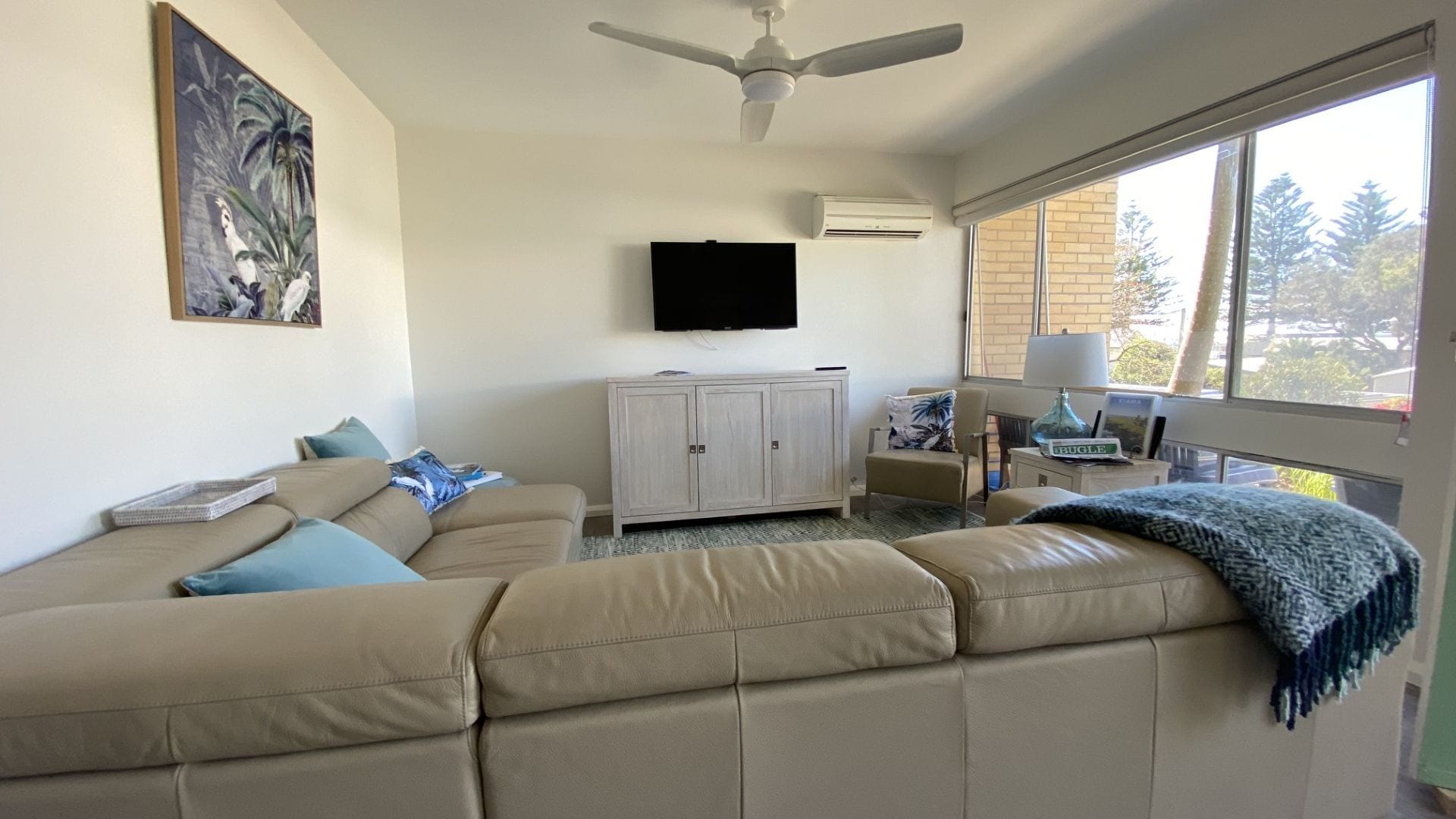 Una sala de estar con un sofá color beige, un mueble, un televisor y un ventanal con vistas a árboles.