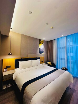 Una habitación de hotel con una cama grande, una mesita de noche con una lámpara, una mesita auxiliar, una ventana con cortinas y una obra de arte colgada en la pared.