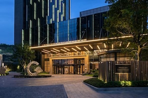 A modern building with a glass facade, a prominent entrance with a canopy, and a landscaped area with a decorative sculpture. - The G Shenzhen, A Tribute Portfolio Hotel (Shenzhen)