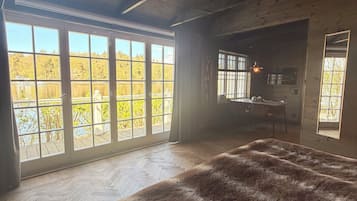 Une chambre dotée d’un grand lit, d’un plancher en bois et offrant une vue sur les arbres grâce à de grandes fenêtres.