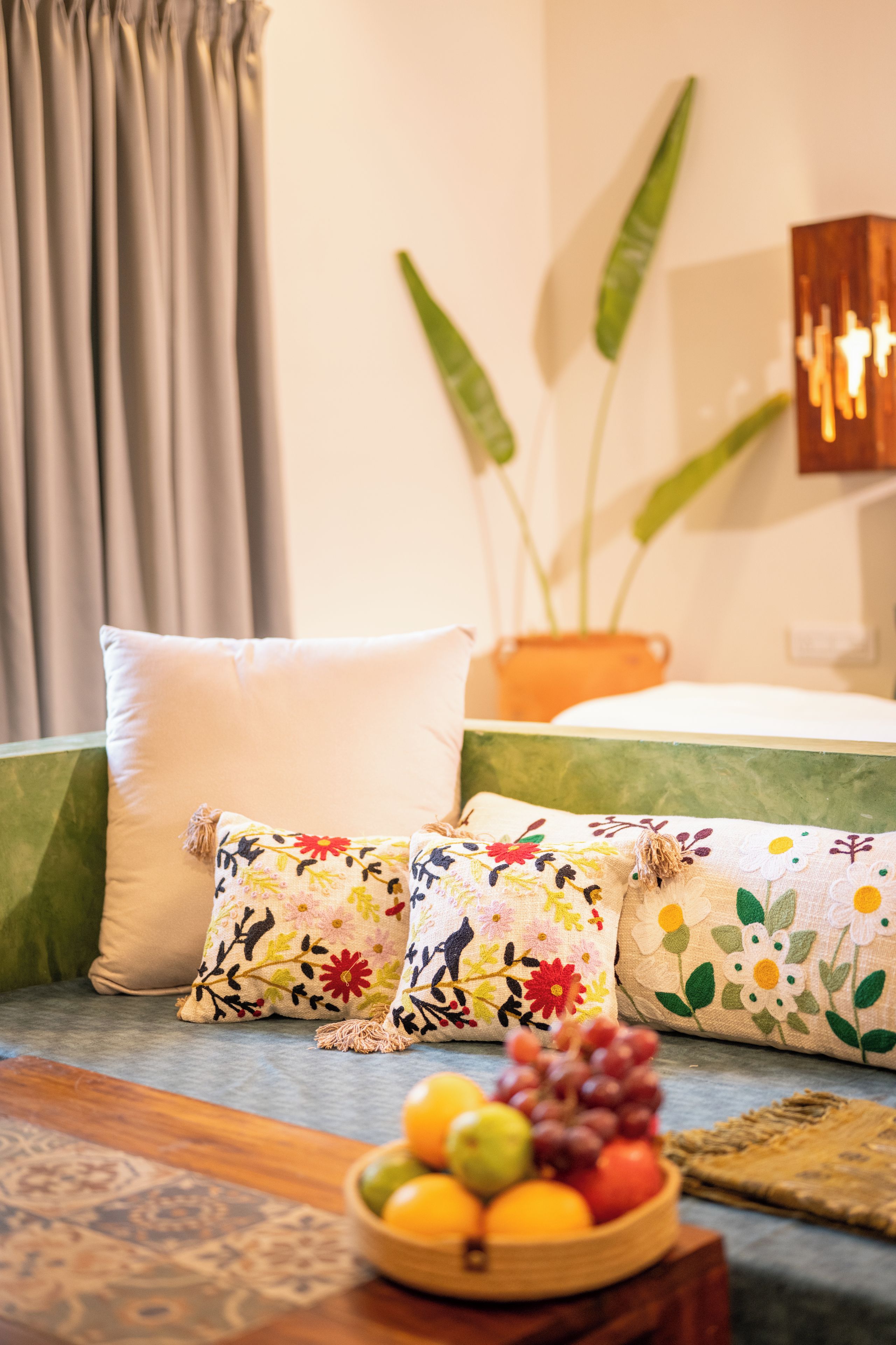 A room with a bed, two pillows, a plant, and a fruit bowl on a table.