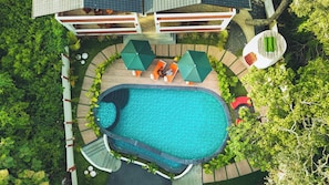 Une vue aérienne d’une piscine entourée de chaises longues et de parasols, le tout bordé d’une végétation luxuriante et d’un bâtiment.