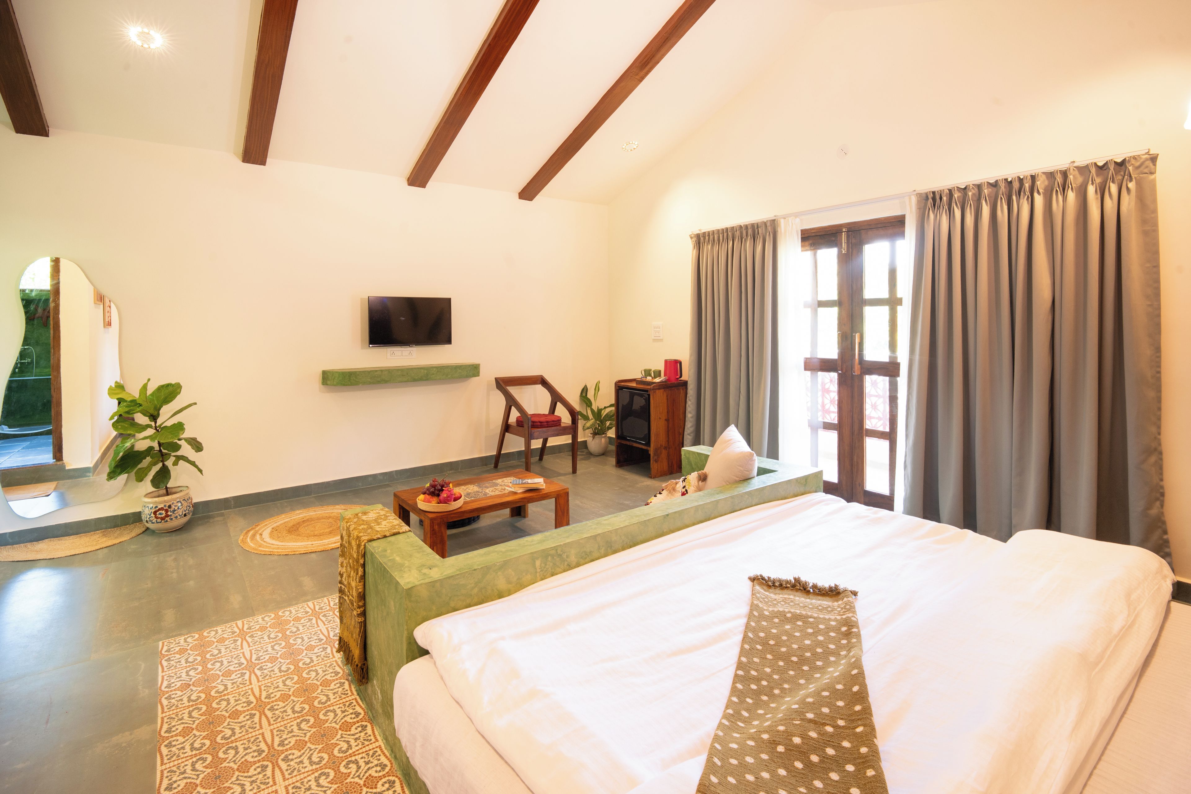 A hotel room with a bed, a television, a desk with a chair, a window with curtains, and a plant.