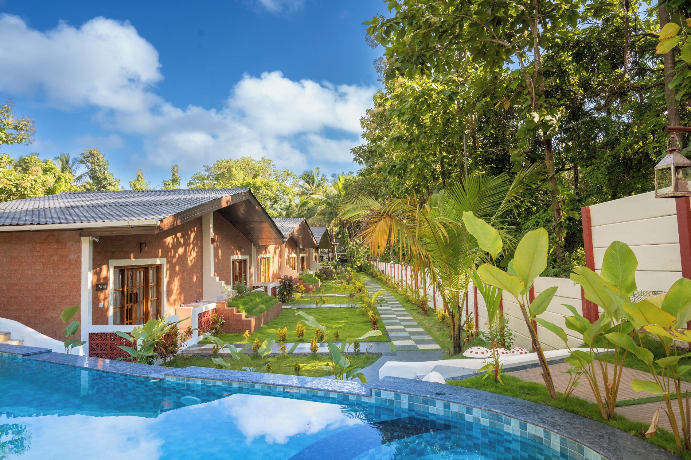 A pool area with a clear sky, a pathway lined with greenery, and a row of buildings with a tiled roof.