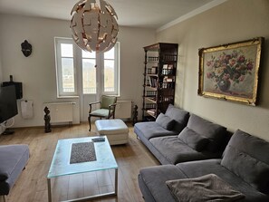 Ein Wohnzimmer mit grauem Sofa, einem Glastisch, einem Holzregal und einem Gemälde an der Wand.