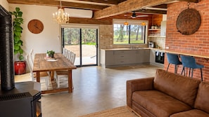 A spacious living area with a wooden dining table, a brown leather sofa, and a kitchen with a brick wall and a large window.