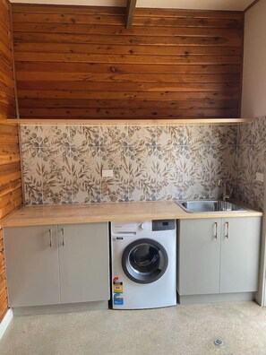 A compact kitchen with a washing machine, wooden ceiling, and tiled backsplash.