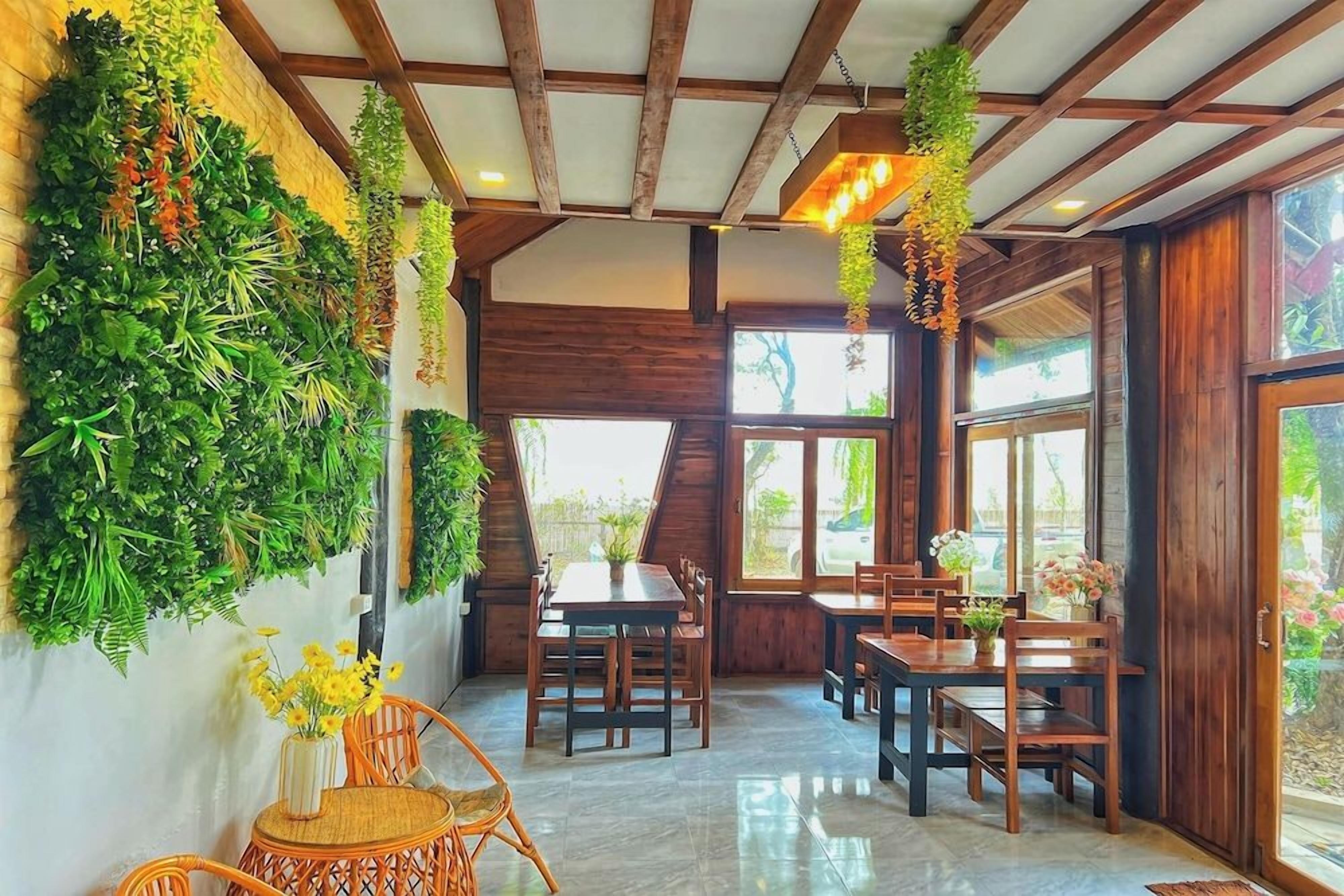 A dining area with wooden tables and chairs, surrounded by lush green walls and hanging plants.