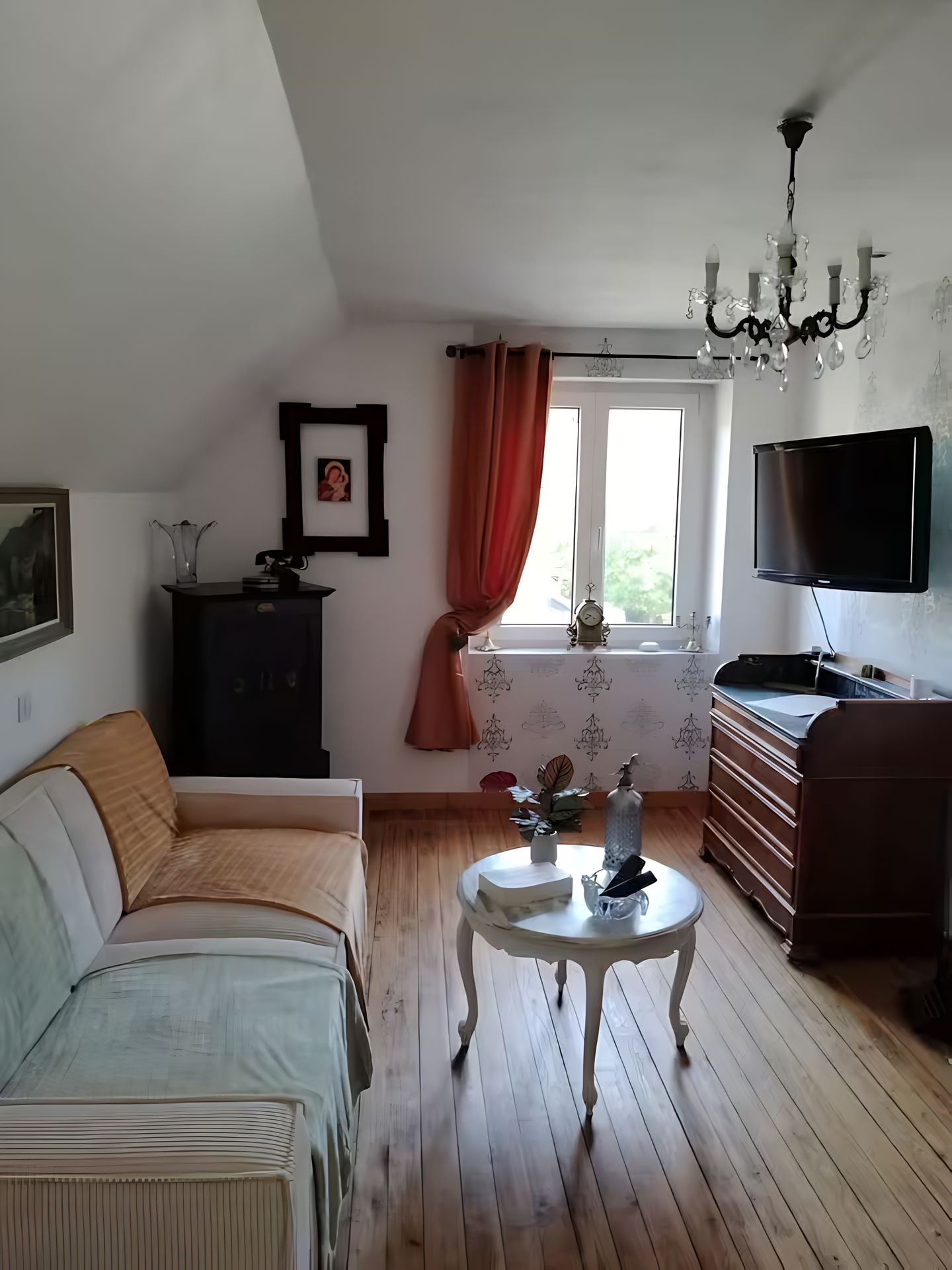 A living room with a sofa, a small table, a television, a chandelier, and a window with curtains.