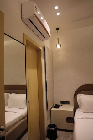 A hotel room with two beds, a bedside table with a phone, a trash bin, and an air conditioning unit. - Varay Prime (Ujjain)