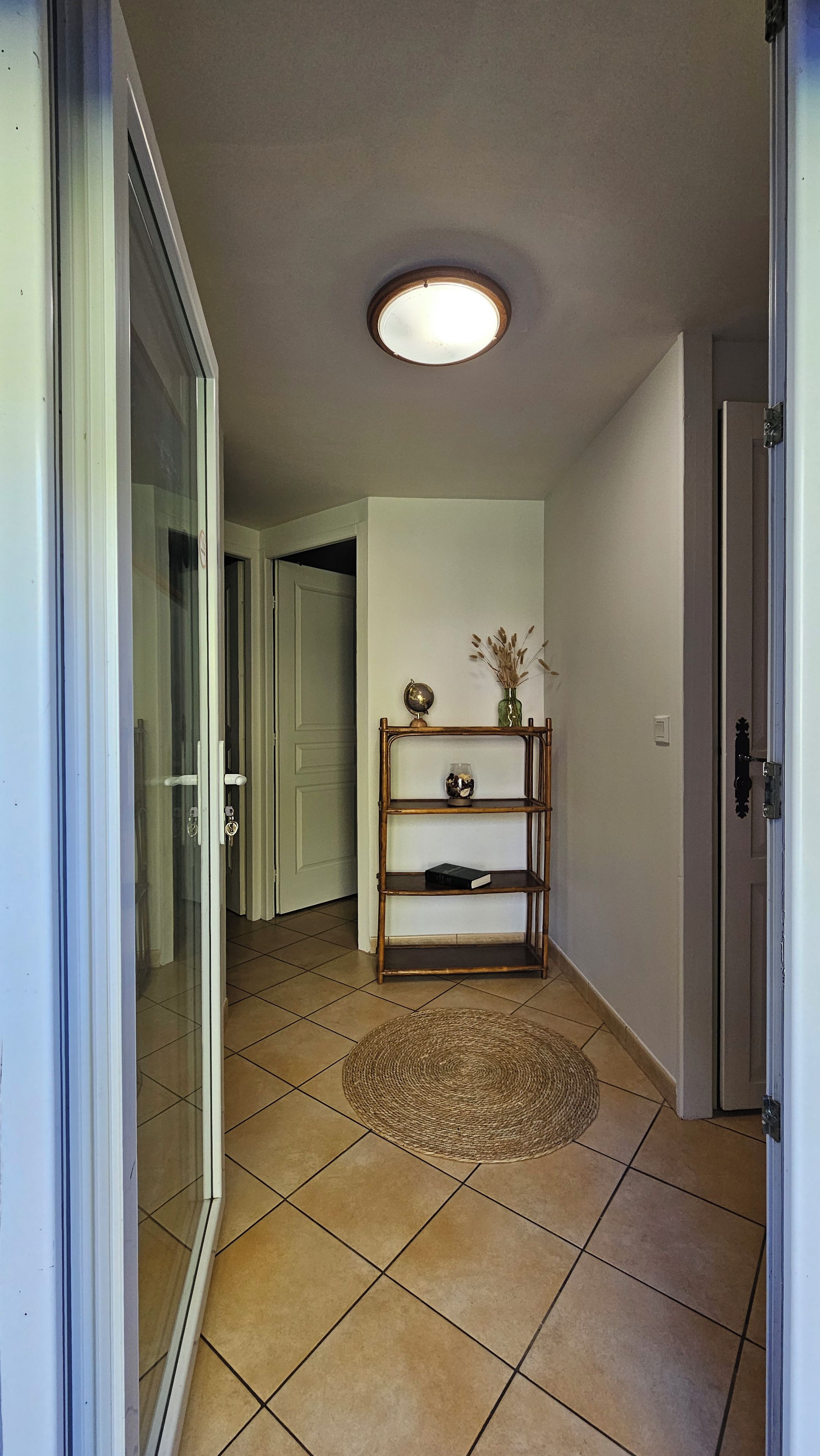 A hallway with a tiled floor, a round woven rug, a wall-mounted shelf with decorative items, and a ceiling light.