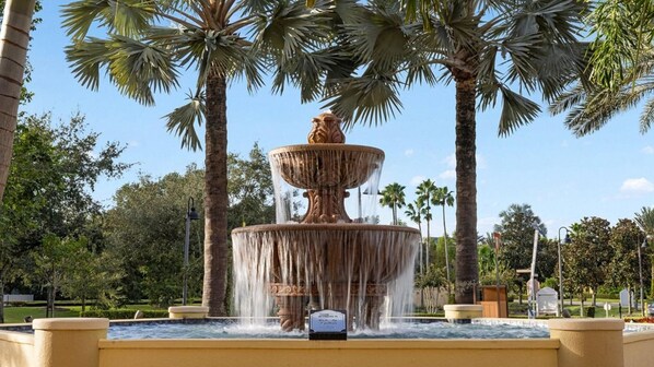 Une fontaine à plusieurs niveaux, dont l’eau jaillit en cascade, entourée de palmiers et surplombée d’un ciel dégagé.