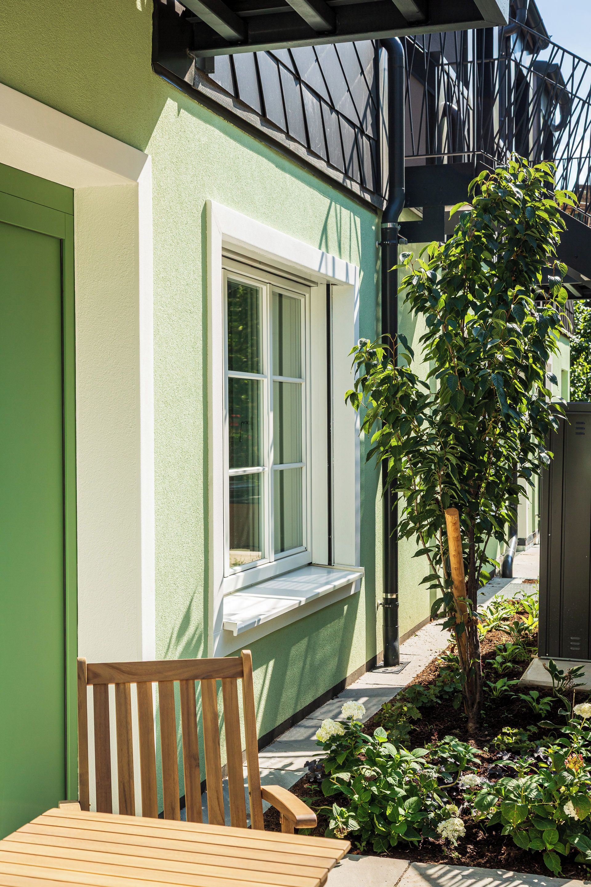 A green building with a window, a wooden bench, and a small tree.