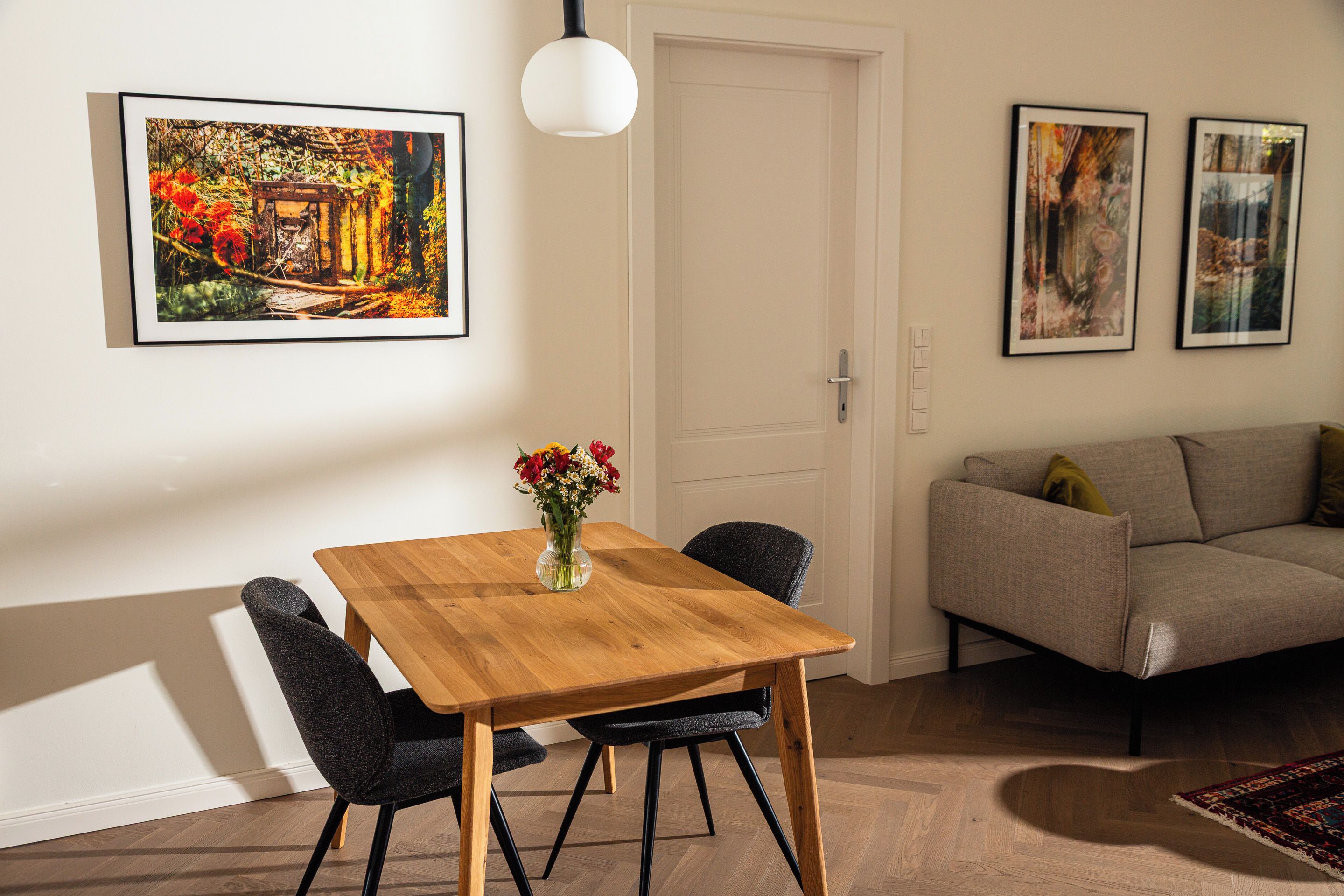 A dining area with a wooden table, chairs, and a vase of flowers. There is a sofa and a door in the background.
