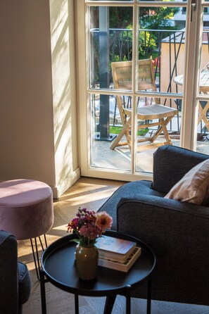 Quarto moderno com sofá, uma mesinha com um vaso e livros, e vista através de uma porta de vidro para uma área externa com mobiliário.