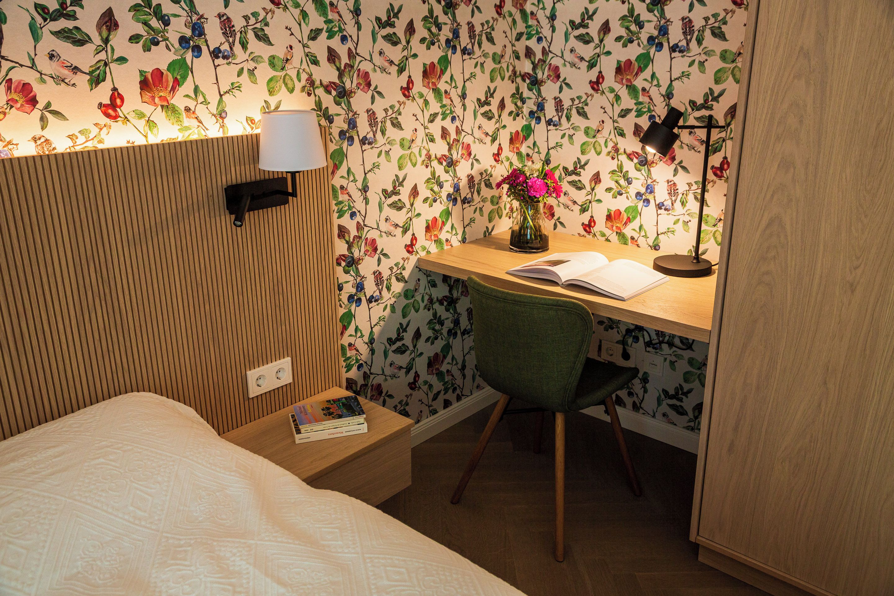 A bedroom with a wooden headboard, a bedside table with a lamp, a green chair, and a desk with a vase of flowers.