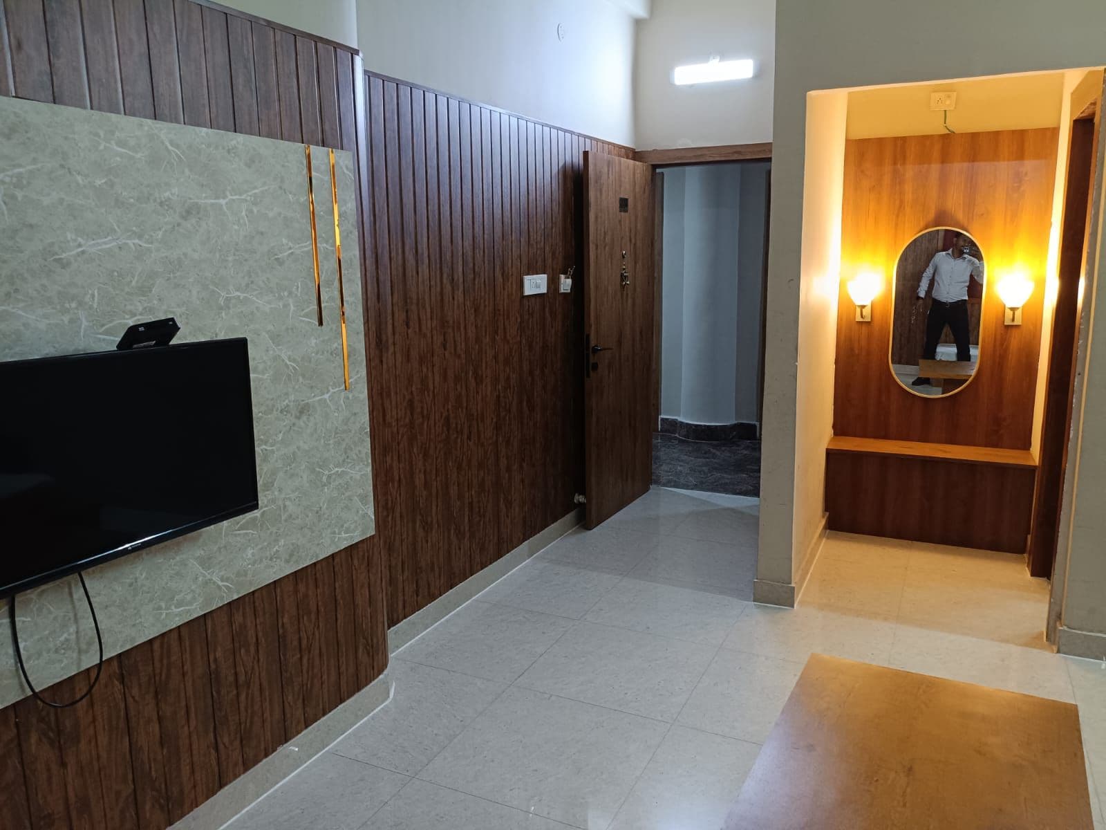 Un pasillo con un televisor de pantalla plana montado en la pared, una puerta de madera y un espejo con apliques.